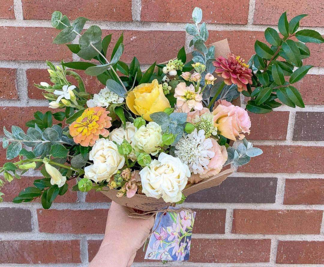 Bouquet of white, gold and peach colored flowers made by Bloom Sacramento held up against a brick wall.