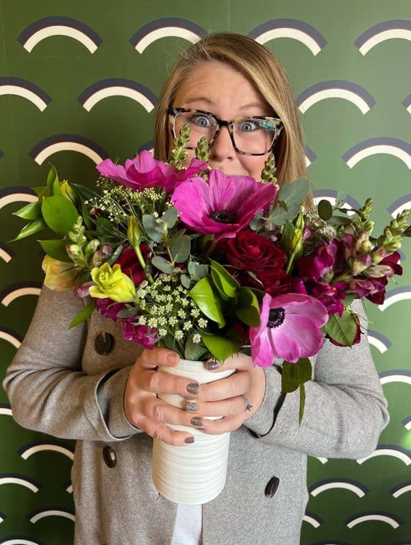 Amanda Kitaura of Bloom Sacramento holds a bouquet of flowers in a white vase, standing in front of a green patterned wall.