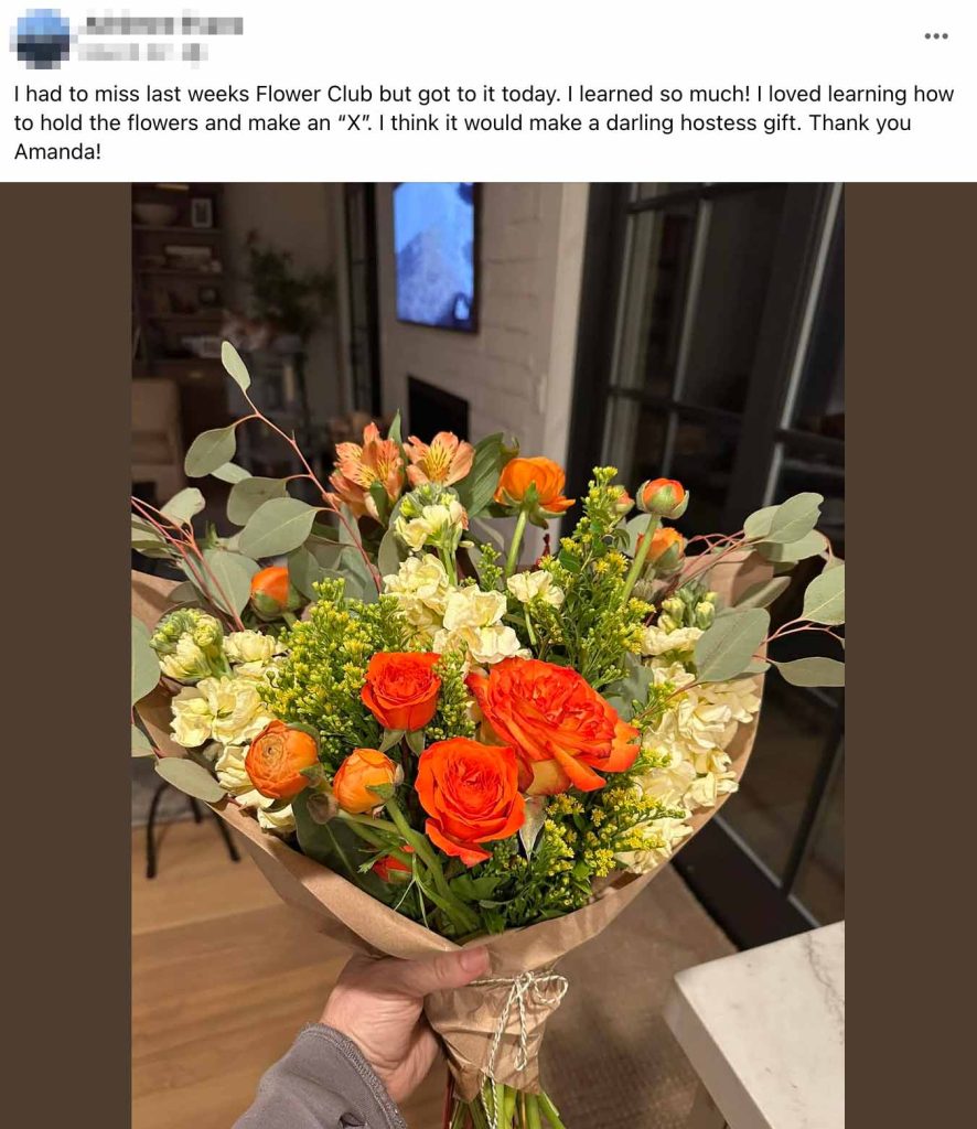 Social media post showing a hand holding an elegant flower arrangement wrapped in brown paper. The bouquet features orange roses, orange ranunculus, cream-colored roses, yellow flowers, and eucalyptus foliage. The post text reads: "I had to miss last weeks Flower Club but got to it today. I learned so much! I loved learning how to hold the flowers and make an 'X'. I think it would make a darling hostess gift. Thank you Amanda!" The photo is taken in a modern living room setting.