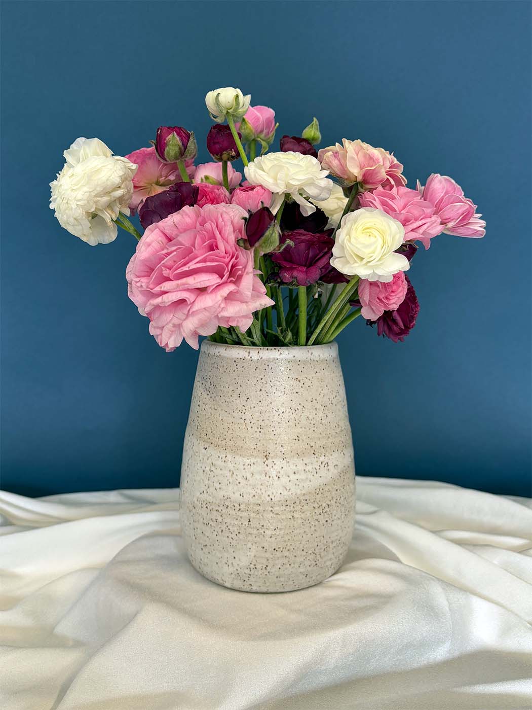 A ceramic vase with flowers sits on a piece of fabric in front of a blue backdrop.